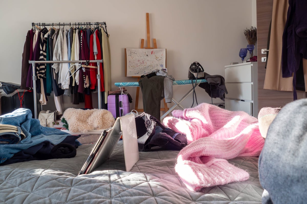 Sunlit bed strewn with clothes, a pink sweater and sneakers among the pillows